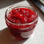 Close-up of a small glass jar filled with vibrant red, textured strawberry freezer jam.