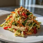 A mound of fresh thai chicken salad featuring shredded chicken, carrots, cabbage, and red peppers, topped with chopped peanuts.